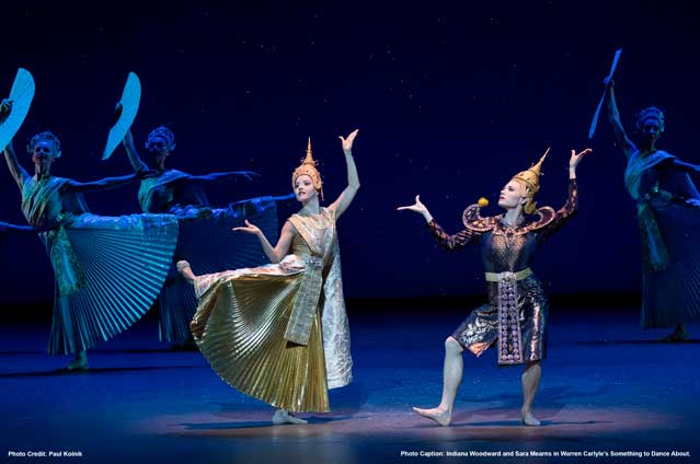 Ballet dancers in ornate, traditional Thai-inspired costumes perform on stage under dramatic lighting; two are in the foreground striking expressive poses, while others hold large, metallic fans in the background.