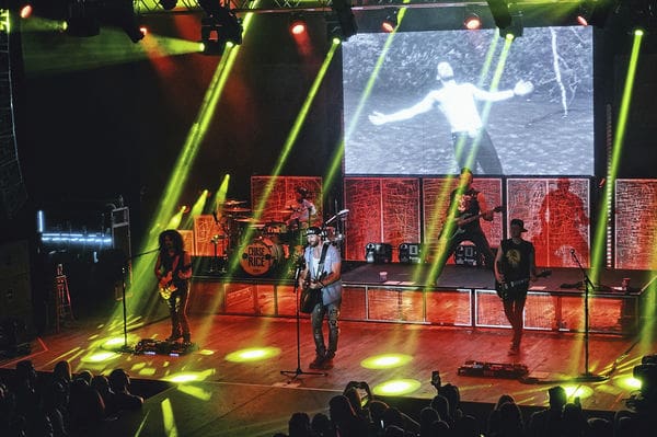 A band performs on stage under yellow spotlights, with dramatic lighting highlighting the drummer, guitarist, and two other musicians. Behind them, a large black-and-white video of a dancing figure plays as the audience watches from below.