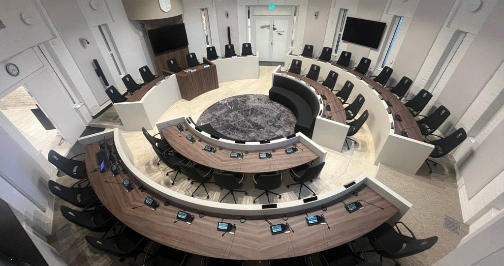 A modern, empty council chamber with curved rows of desks and black chairs arranged in a semicircle around a central circular carpet, featuring advanced lighting, microphones, and digital screens at each seat.