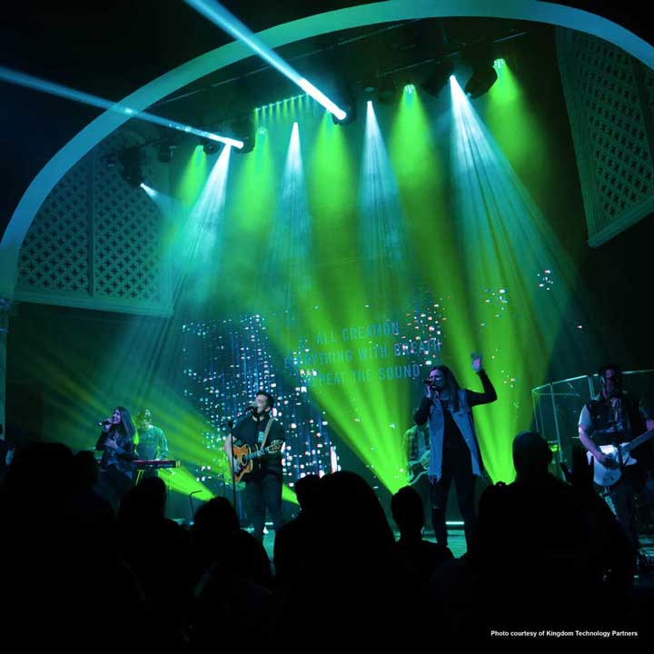A band performs on stage under vibrant green and blue lighting, with musicians playing guitar, keyboard, and singing, while an audience watches in silhouette.