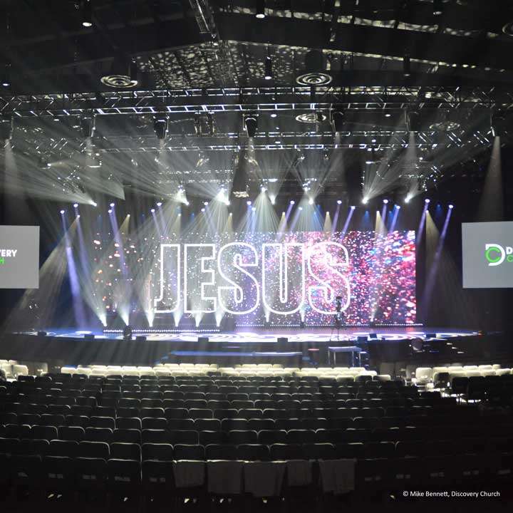 A large indoor stage with bright audio and lighting, plus a screen displaying the word JESUS in bold letters, faces rows of empty chairs in an auditorium setting.