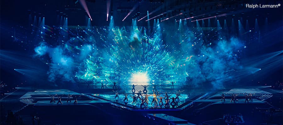A large stage is lit with vibrant blue and green lighting, creating an energetic, cosmic effect. Numerous performers in silhouette pose dramatically in front of the illuminated backdrop as audio pulses and the audience is dimly visible.