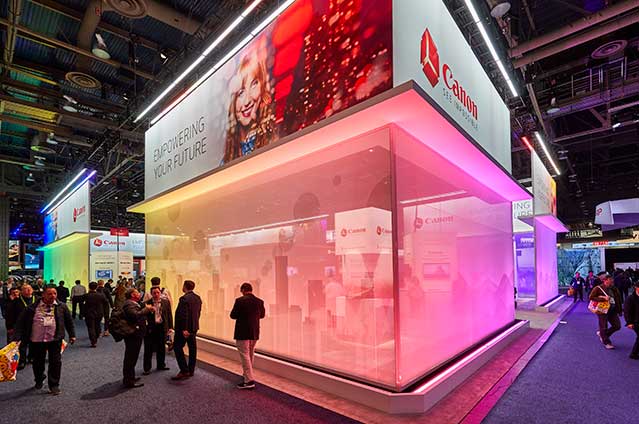 A brightly lit Canon booth at a tech convention, featuring vibrant lighting panels, people walking by, and a large sign displaying the Canon logo with the slogan Empowering Your Future.