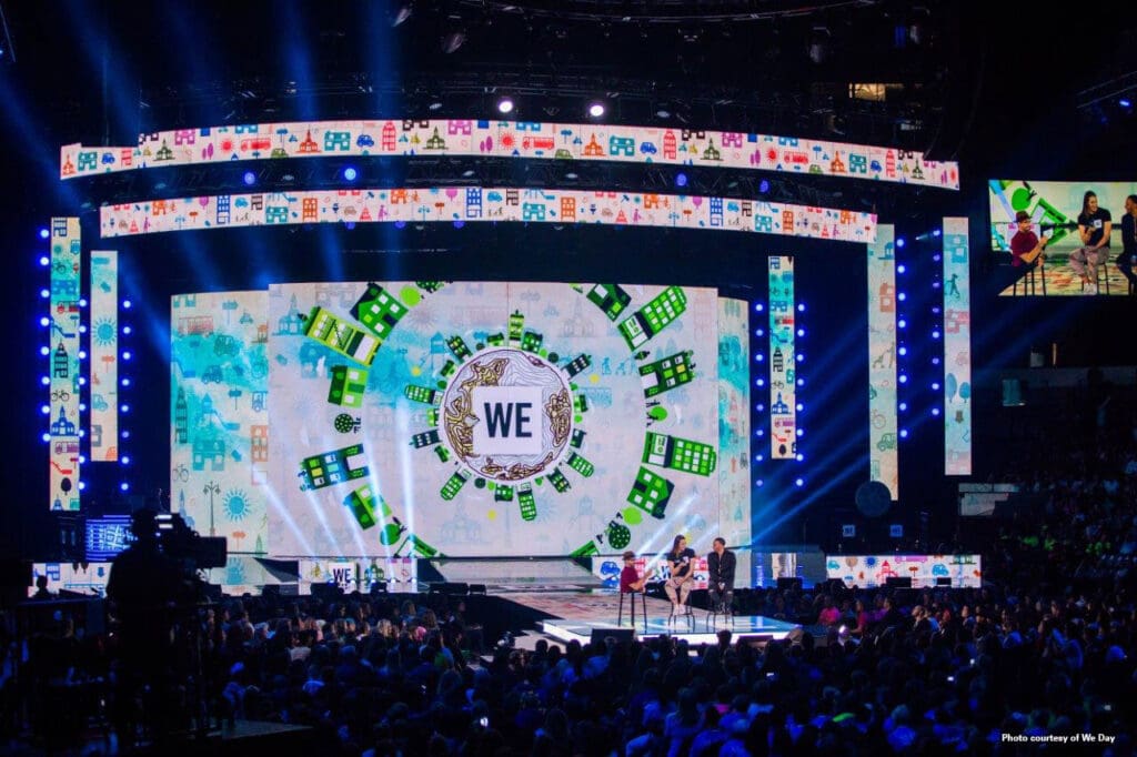 A large crowd faces a brightly lit stage with colorful graphics and the word WE at the center, as speakers stand on stage. Dynamic lighting and video screens surround the arena, creating an energetic event atmosphere.