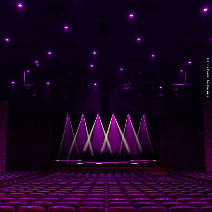 An empty theater with rows of red seats faces a stage lit by purple spotlights, casting beams upward. Subtle purple lighting enhances the dramatic atmosphere, while the setup promises an immersive video and audio experience.