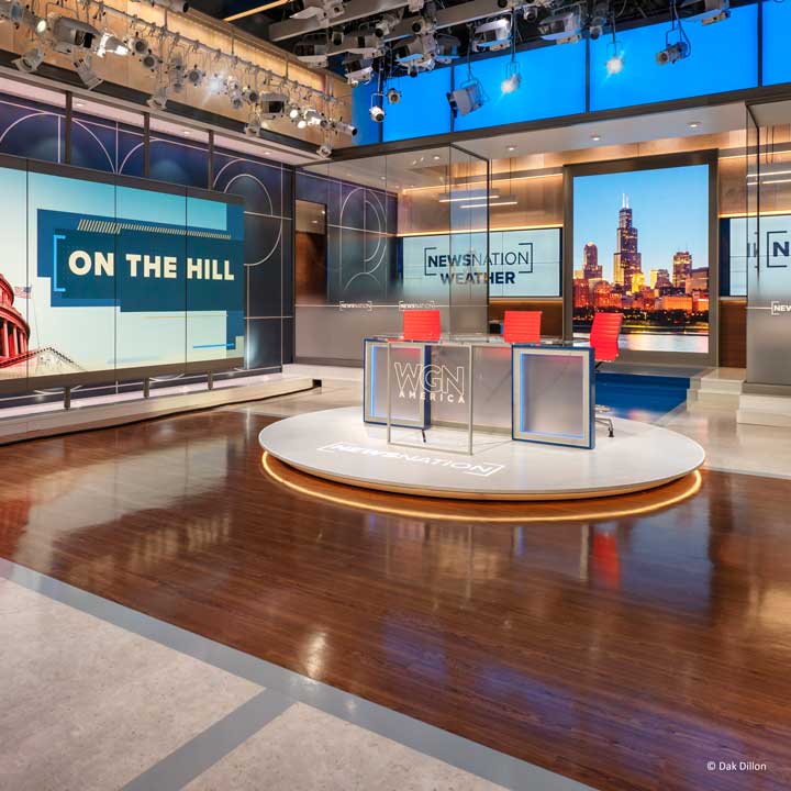 A brightly lit television news studio features three red chairs at a round desk, large digital screens displaying “ON THE HILL” and “NewsNation Weather,” excellent audio setup, and a cityscape graphic in the background.