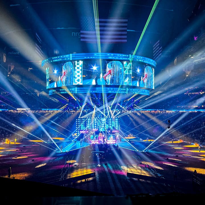 A vibrant concert scene in a packed arena with colorful lighting effects, a large circular video screen above the stage, and a cheering audience surrounding the performers below.