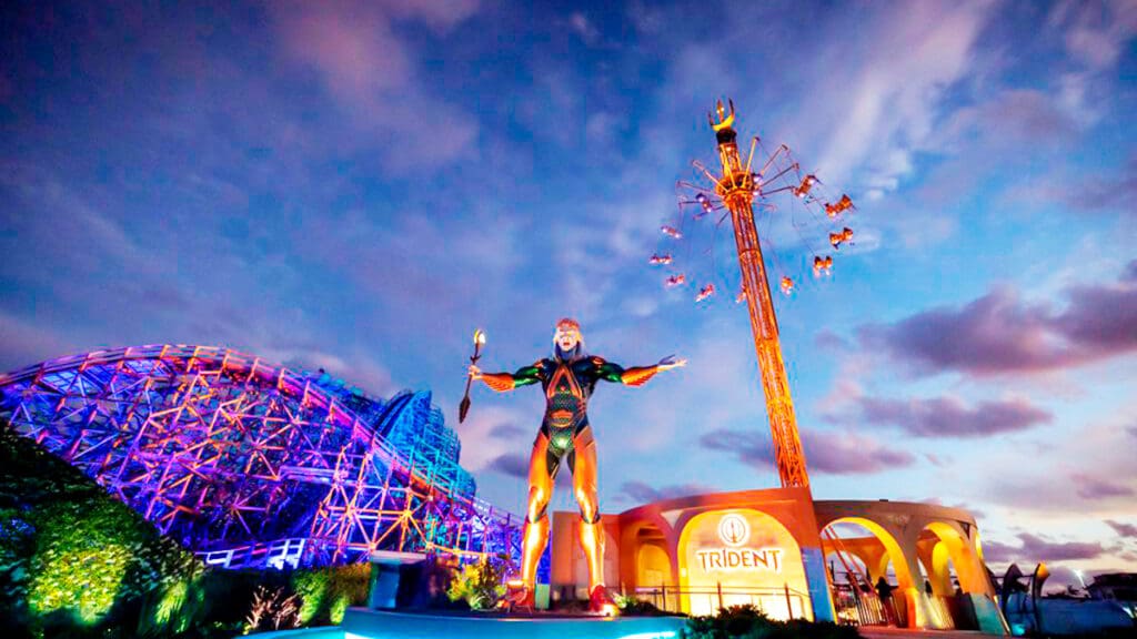 A large statue of Poseidon holding a trident stands in front of a colorful roller coaster and a tall spinning ride called Trident, set against a dramatic, partly cloudy evening sky.