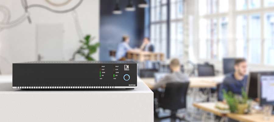 A black network device with indicator lights sits on a white surface in the foreground; in the background, people work at desks under modern office lighting in a bright, contemporary workspace.
