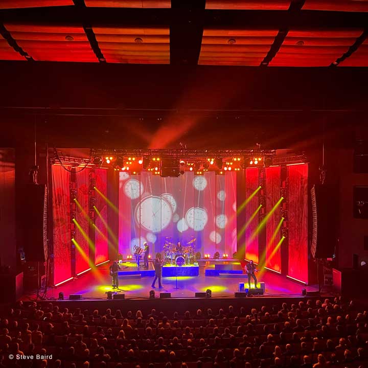 A band performs on a brightly lit stage with dynamic red and yellow lighting and clock graphics projected on the video backdrop, as an audience watches in a darkened theater.