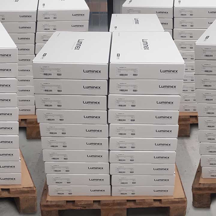 Stacks of white boxes labeled Luminex—used for audio, video, and lighting equipment—are arranged on wooden pallets in a warehouse. Each box has barcodes and printed labels, all neatly organized in several tall columns.