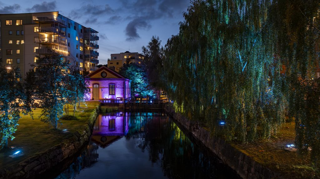A canal at dusk reflects nearby trees and buildings. An old brick building is lit with colorful purple and blue lights, and modern apartment blocks are visible in the background among illuminated greenery. A project by LumenRadio
