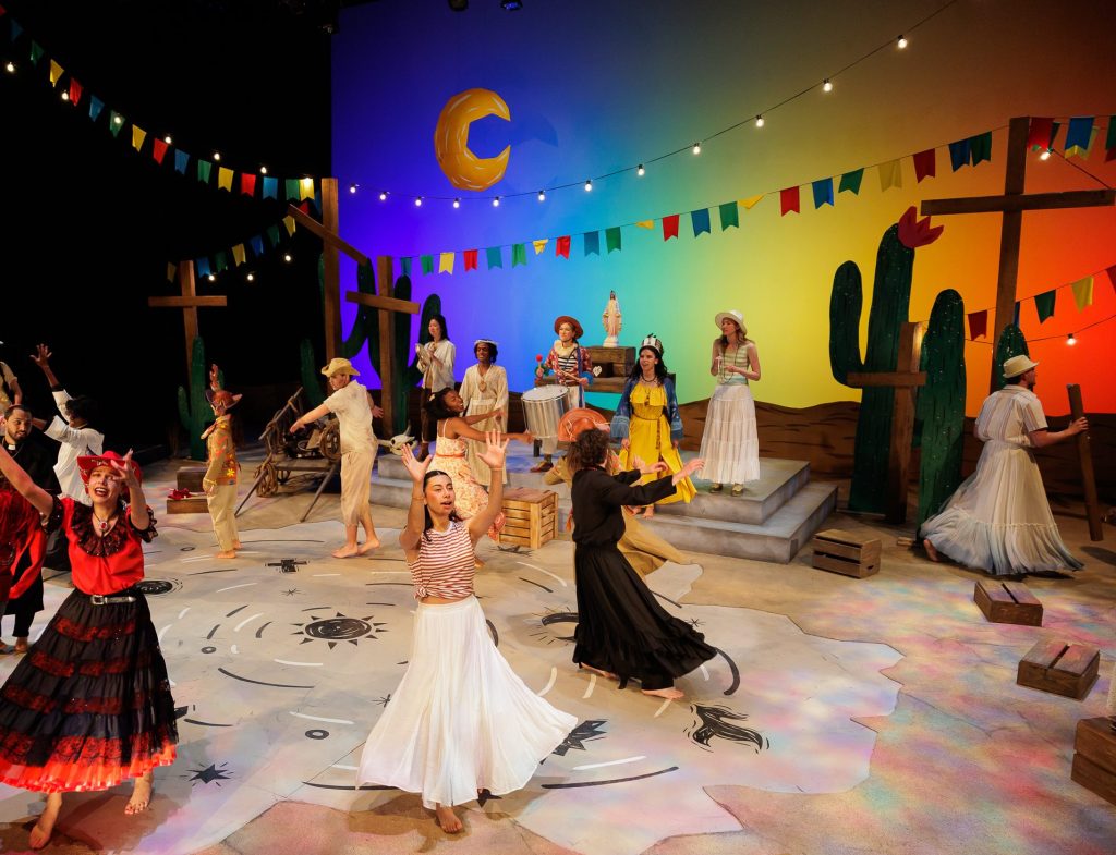 A lively theater scene shows performers dancing in colorful costumes on a set decorated with cacti, crosses, bunting, and a painted backdrop of a crescent moon and sunset, evoking a festive desert atmosphere.