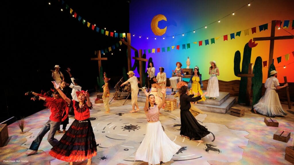 A vibrant stage scene with actors in colorful costumes dancing and singing. The set features wooden crosses, cacti, festive string flags, and a painted sunset backdrop with a crescent moon. The mood is lively and celebratory. (c) Rich Soublet