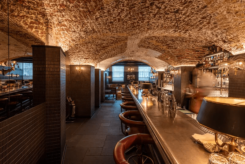 A cozy bar with a vaulted brick ceiling, dim lighting, leather bar stools, and a long counter. Shelves with bottles line the walls, and a blurred bartender moves behind the bar.