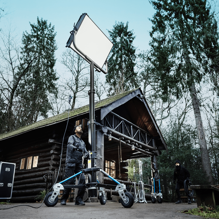 A person adjusts a large studio lighting fixture on a wheeled stand outside a rustic wooden cabin surrounded by tall trees, with another person and video equipment visible in the background.
