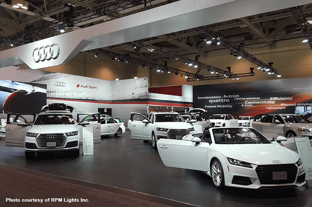 Audi car display at an auto show, featuring several white Audi vehicles on a well-lit showroom floor with dramatic lighting, informational signs, and branded banners overhead.
