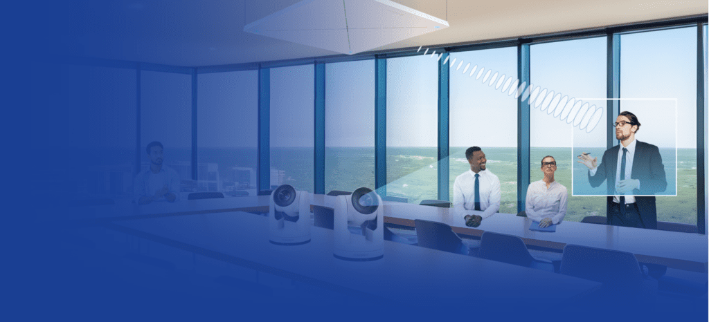 Three people sit at a conference table in a glass-walled office overlooking the ocean, while a presenter stands and gestures toward a whiteboard. Conference cameras and audio equipment are on the table.