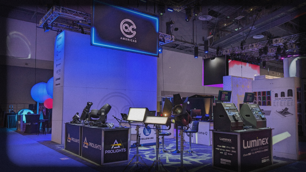 A booth at a trade show displays professional AVL equipment with spotlights and LED fixtures on stands. The booth has branding for AC Americas, with overhead screens and banners.