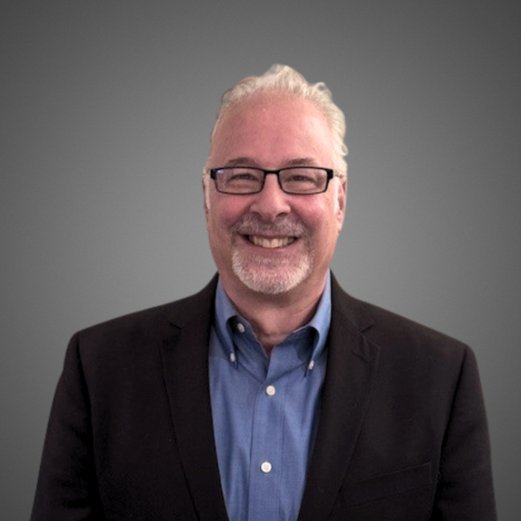 Smiling middle-aged man with short white hair, glasses, and a beard, wearing a dark blazer over a blue shirt, standing in front of a plain gray background.