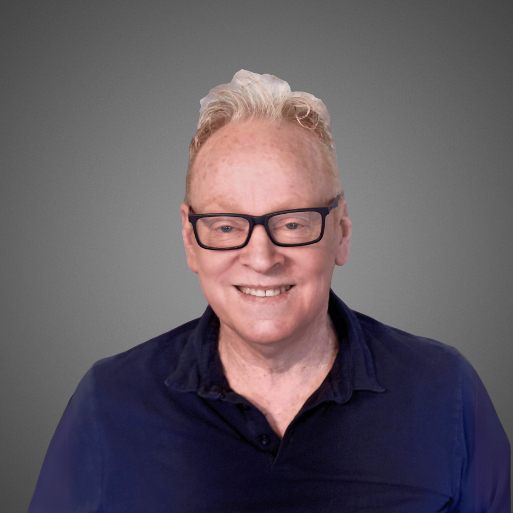 A smiling older man with light skin, short white hair, and black glasses wearing a dark blue collared shirt poses against a plain gray background.