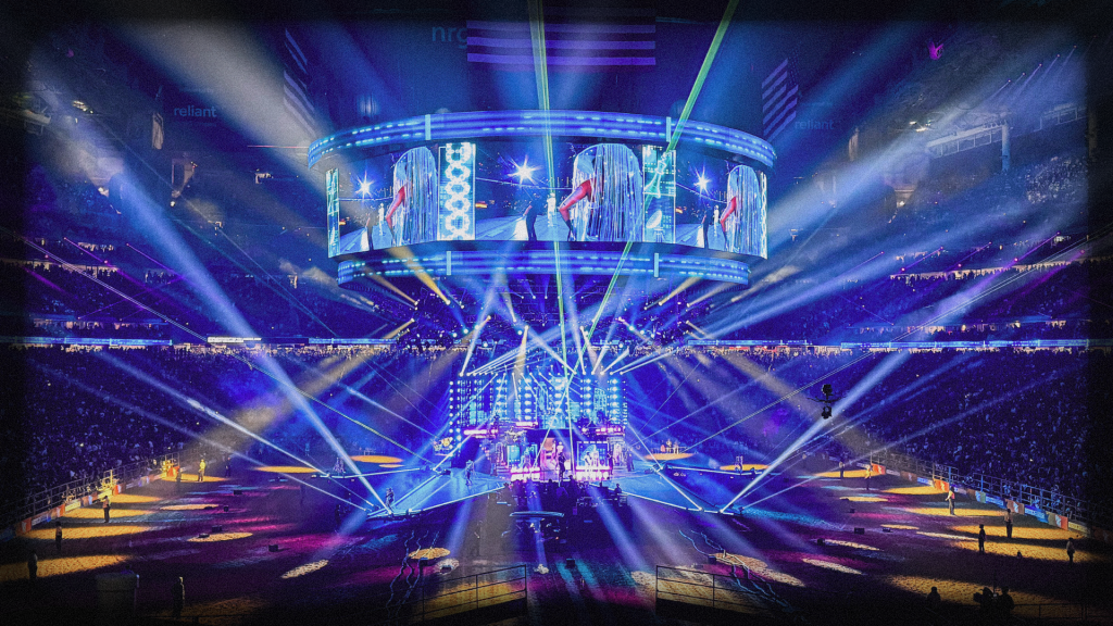 A concert stage in a packed stadium is illuminated by colorful spotlights and lasers, with a large circular screen above showing performers. The audience surrounds the vibrant, high-energy scene.