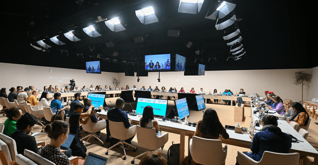 A large group of people sit around a circular conference table in a well-lit meeting room, with multiple screens, microphones, and advanced audio setup, participating in a formal discussion or conference.