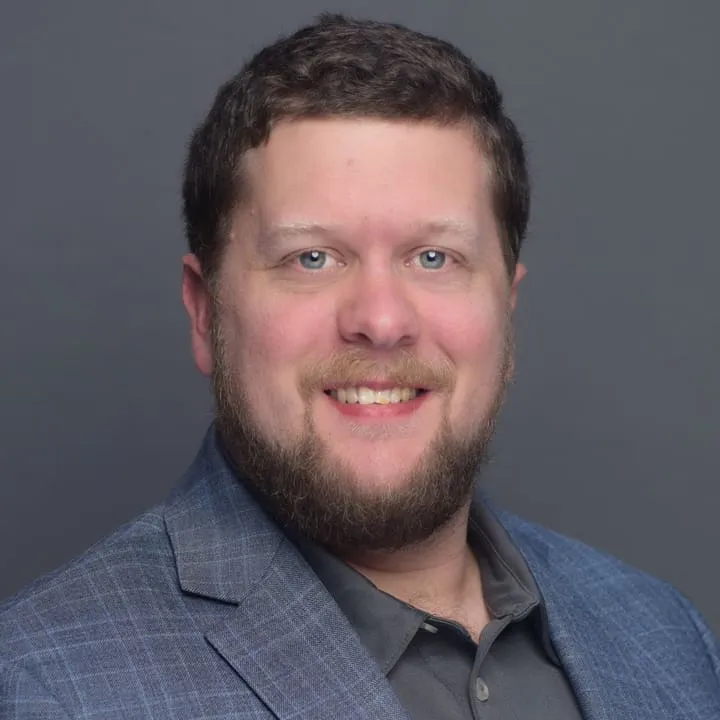 A man with a beard and short brown hair, wearing a blue checked blazer over a gray collared shirt, smiles in front of a plain gray background with soft lighting highlighting his features.