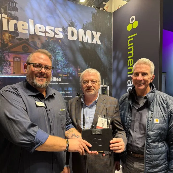 Three men stand smiling in a convention booth with a Wireless DMX sign and lumenea branding in the background. Two of them are holding a black electronic lighting device together.