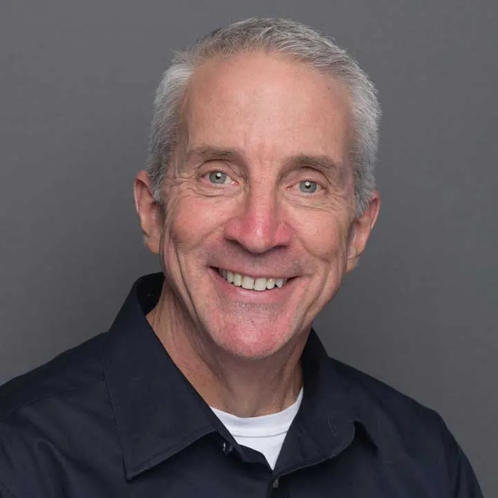 Smiling older man with short gray hair wearing a dark collared shirt and white undershirt, posing against a plain gray background with professional lighting.